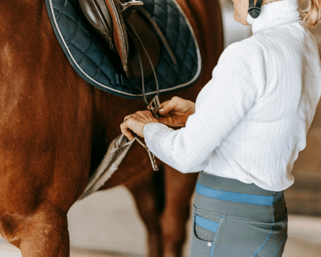 Close-up of a woman tightening an English saddle on a chestnut horse, representing preparation, tradition, and the care behind every ride.
